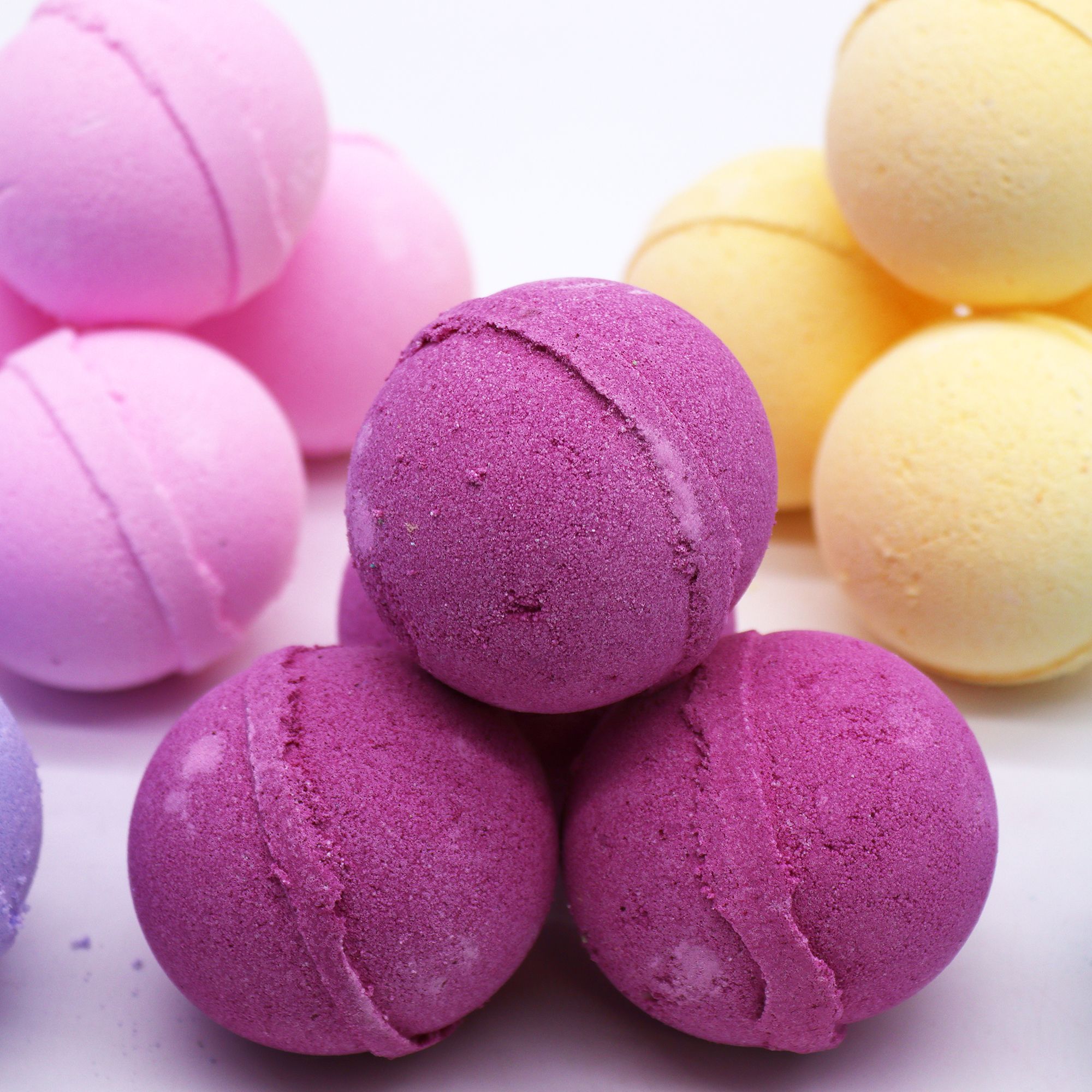 Tres bombas de baño moradas en primer plano con otras bombas de colores al fondo correspondiente a bomba de baño ylang ylang y jengibre.