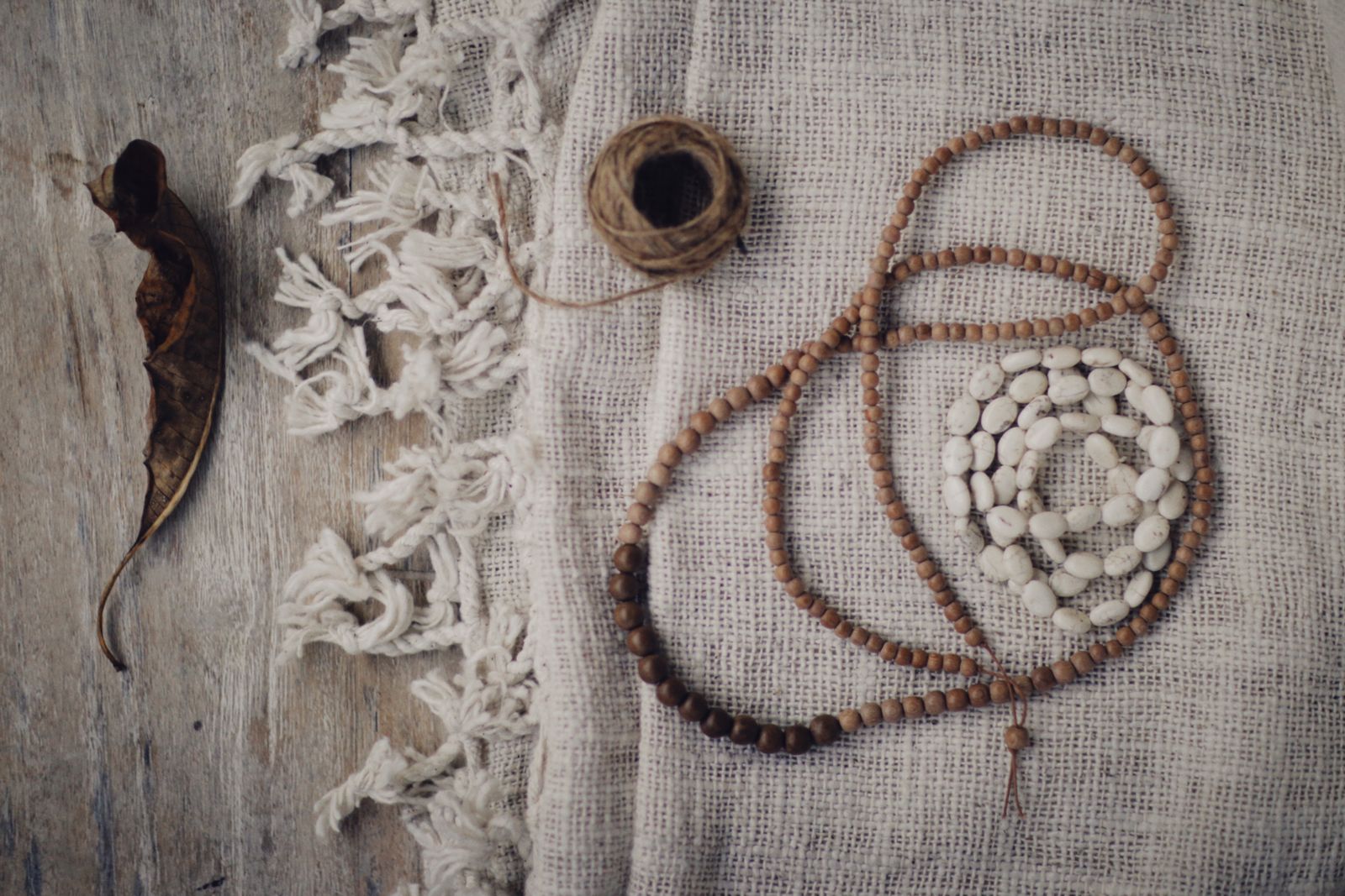 Collar largo de cuentas de madera y cuentas blancas de piedra, junto a un ovillo de cordel de yute, extendido sobre tela de lino crudo y madera desgastada, visto desde arriba