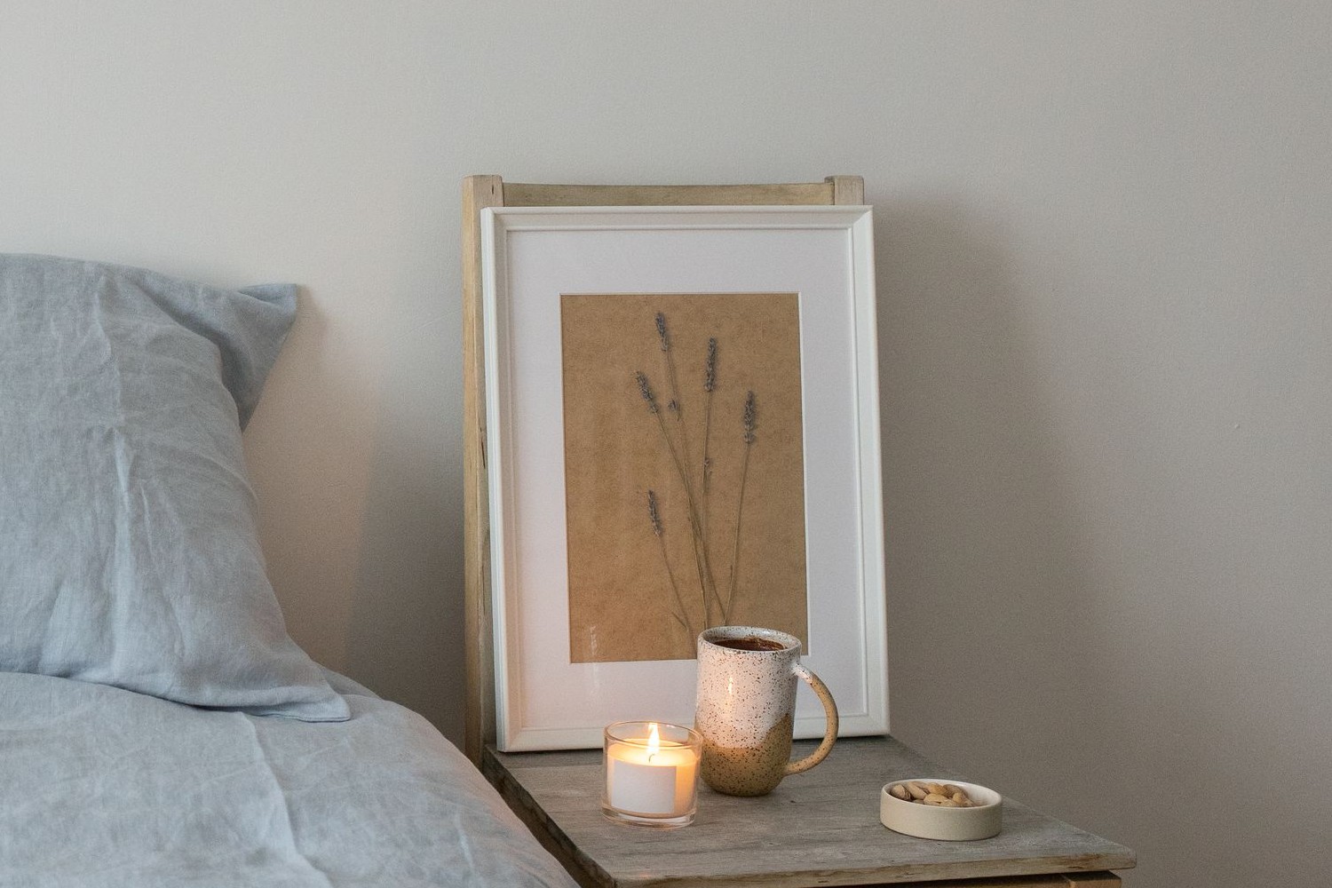 Mesilla de noche de madera junto a una cama vestida de lino gris azulado, con una vela encendida en vaso, una jarra de cerámica moteada y una lámina enmarcada de espigas de lavanda secas apoyada en la pared beige
