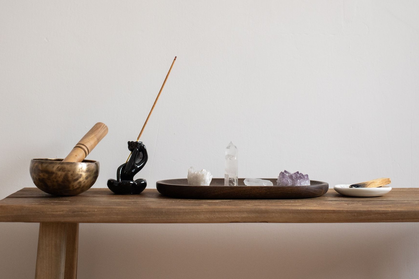 Cuenco tibetano de metal con maza de madera, portaincienso negro con una varilla y bandeja ovalada de madera con puntas de cuarzo transparente, una drusa de amatista y palo santo, sobre un banco de madera clara ante una pared beige