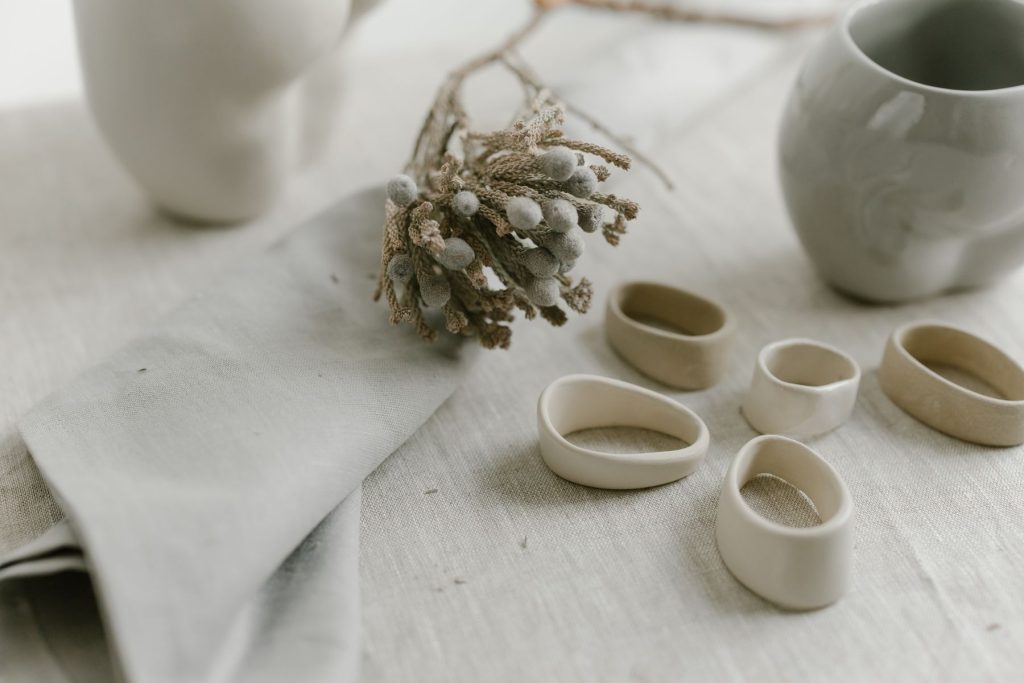 Bodegón cercano sobre un mantel de lino crudo: una ramita de flores secas de color beige apoyada en una servilleta doblada, varios aros de cerámica artesanal en tonos crema y, al fondo desenfocados, una jarra y un vaso de cerámica clara