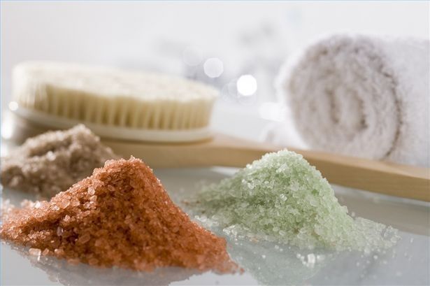 Dos montones de sales de baño de distintos colores junto a un cepillo y una toalla.